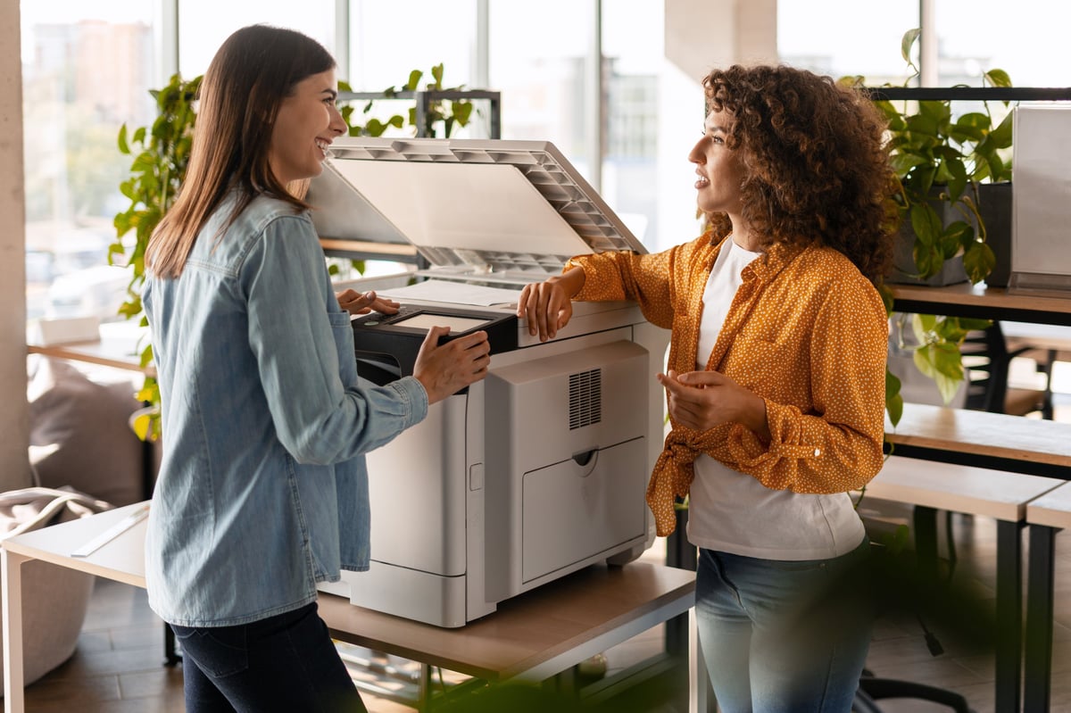 Two young businesswomen smiling while collaborating at a multifunctional printer in a sleek, modern office environment