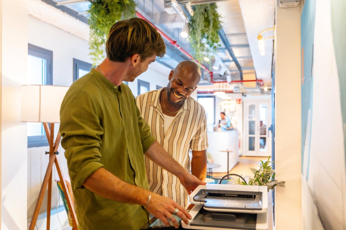 Two colleagues laugh together by the office printer, sharing a lighthearted moment of teamwork and connection in a modern, casual workplace environment