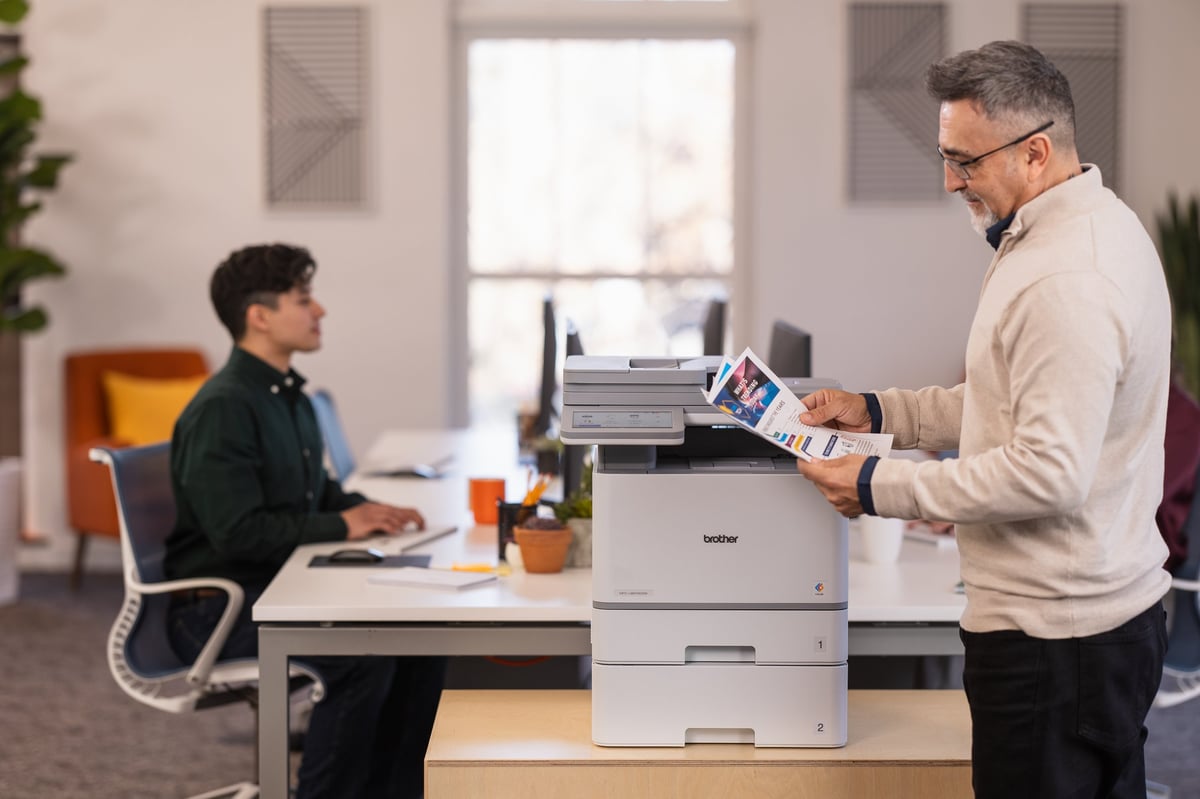 Man holding printed documents next to office printer with colleague seated at desk in modern workplace